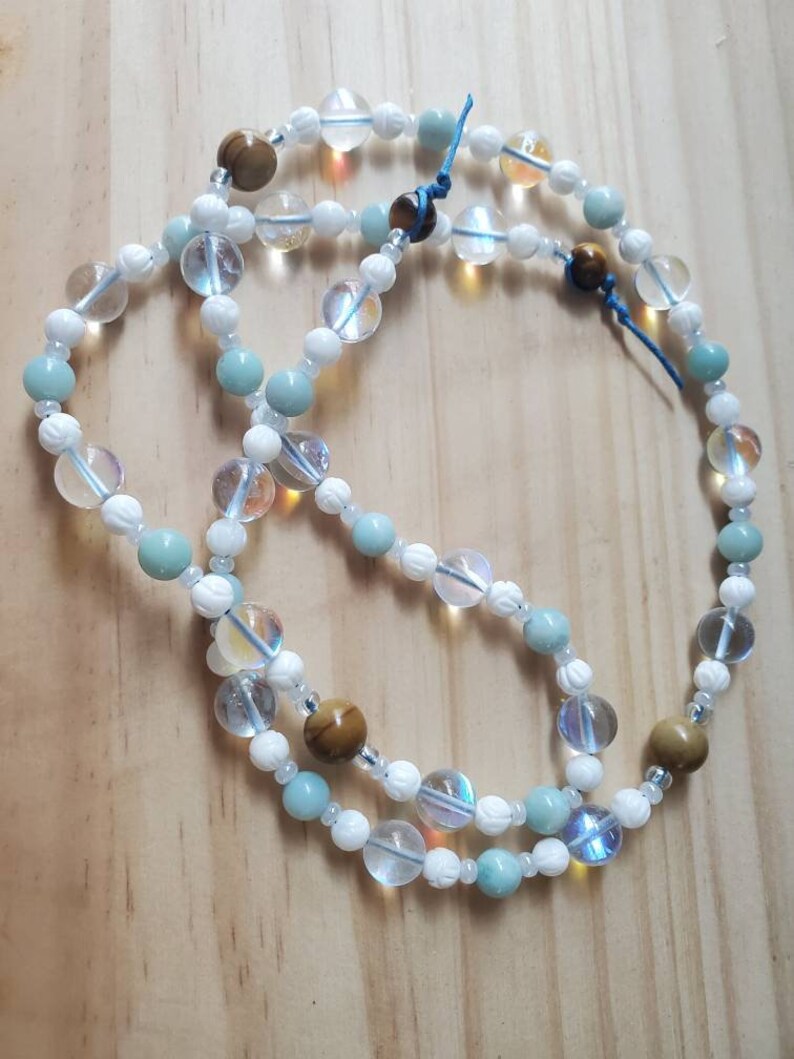 May include: A multi-strand necklace with white, light blue, and brown beads. The necklace is made of various types of stones, including clear quartz, aquamarine, and tiger's eye.