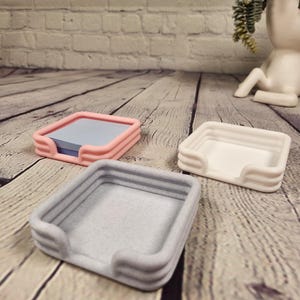May include: Three square desk organizers in pink, white, and gray, arranged on a wooden surface. The pink organizer holds a stack of blue notepaper. The organizers have a minimalist design with rounded edges and a textured appearance.