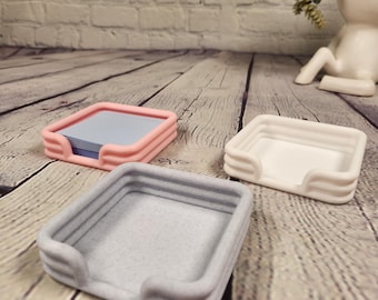 May include: Three square desk organizers in pink, white, and gray, arranged on a wooden surface. The pink organizer holds a stack of blue notepaper. The organizers have a minimalist design with rounded edges and a textured appearance.