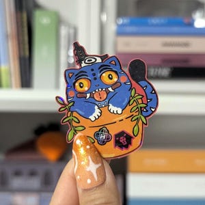 May include: A blue cat sticker with a unique design, featuring a cat with yellow eyes, a red tongue, and a black tail. The cat is inside an orange container with green leaves. The sticker has a holographic effect.