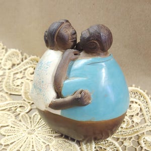 Puede incluir: Una escultura de cerámica de dos figuras abrazándose. Una figura lleva una camisa azul claro, la otra una camisa blanca. La escultura tiene una base marrón y cabezas marrones oscuras. Las figuras están en un abrazo amoroso.