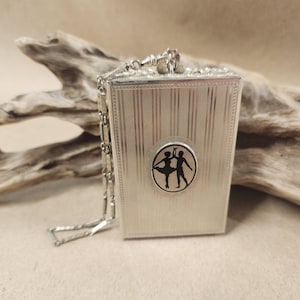 May include: A silver-toned rectangular compact with a chain. The front features a circular emblem with black silhouettes of two dancers. The compact has vertical striped detailing and a decorative clasp.