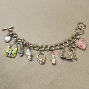 Puede incluir: Una pulsera de dijes plateada con varios dijes colgantes. Los dijes incluyen un bolso, un tacón alto, un reloj, un anillo, un lápiz labial, gafas de sol, un corazón y llaves. La pulsera tiene un cierre de palanca y un diseño de eslabones de cadena.