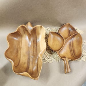 Puede incluir: Dos cuencos de madera para servir. Uno es un cuenco grande, de forma irregular, con un borde ondulado. El otro es un cuenco más pequeño, con forma de hoja y cuatro compartimentos. Ambos cuencos están hechos de madera marrón claro y tienen vetas naturales.