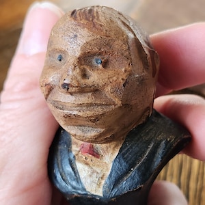 Op de afbeelding: Een gesneden houten buste van een man met een bruin gezicht, blauwe ogen en een zwart pak. De buste heeft een gedetailleerd, getextureerd uiterlijk, met zichtbare snijtekens. Een rode stropdas is zichtbaar. De buste wordt in een hand gehouden.