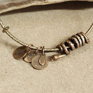 May include: A gold-coloured metal bracelet featuring a coiled snake design and three small, round charms. The snake's head is detailed, and the charms have etched designs and text. The bracelet is on a neutral surface.