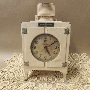 Puede incluir: Un reloj antiguo de color blanquecino con un cuerpo cuadrado y una esfera redonda. La esfera del reloj tiene números y manecillas negras, con las palabras "Edison" y "General Electric" visibles. El reloj tiene una parte superior cilíndrica y se apoya en cuatro patas pequeñas.