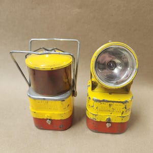 May include: Two vintage Justrite lanterns. One is a lantern with a handle and a yellow top and base, a brown midsection, and a red bottom. The other is a headlamp with a large lens and a yellow and red body.