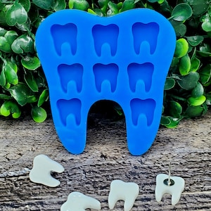 May include: Blue silicone mold shaped like a tooth with nine individual tooth-shaped cavities.  There are four white tooth-shaped resin pieces on a wooden surface.