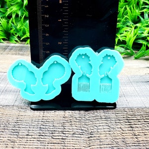 May include: A turquoise blue silicone mold with four cavities. Two cavities are shaped like a woman's head with an afro hairstyle. The other two cavities are shaped like a comb.
