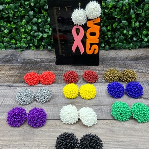 May include: Assortment of beaded earrings in various colors, including red, silver, yellow, white, purple, green, blue, and black. The earrings are displayed on a wooden surface.