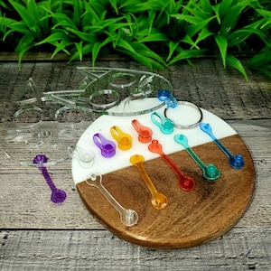 May include: A set of colorful acrylic keychain charms in the shape of a cat, with a rainbow of colors including purple, blue, green, yellow, orange, and red. The charms are displayed on a wooden and white circular base.