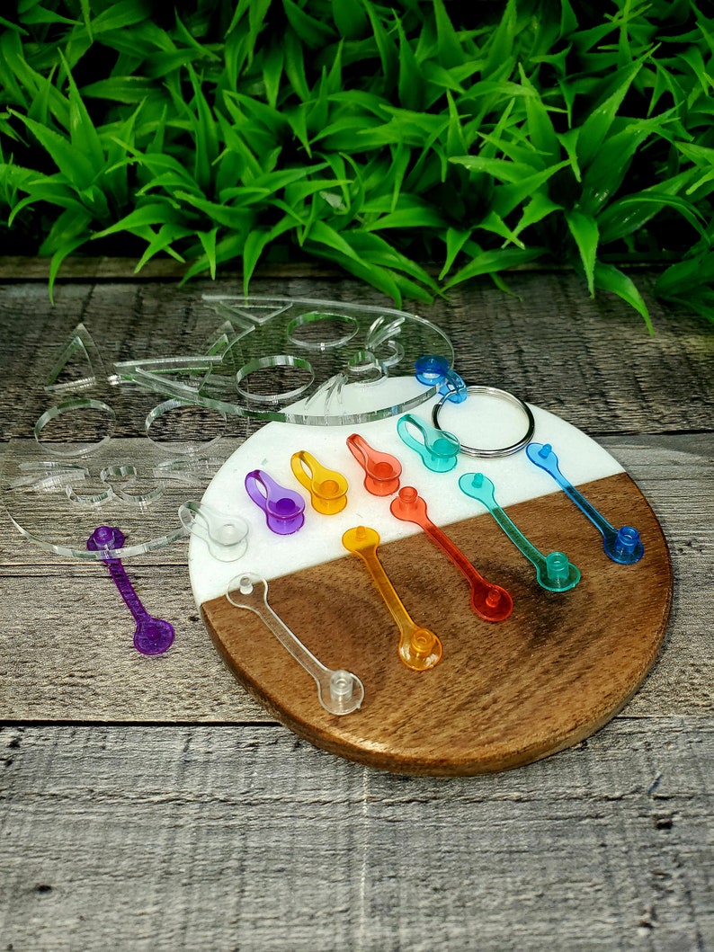 May include: A set of colorful acrylic keychain charms in the shape of a cat, with a rainbow of colors including purple, blue, green, yellow, orange, and red. The charms are displayed on a wooden and white circular base.