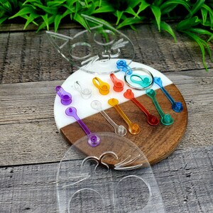 May include: A set of colorful plastic keychains in the shape of a cat. The keychains are arranged on a wooden and white circular board. The keychains are in various colors, including purple, blue, green, orange, yellow, and red.