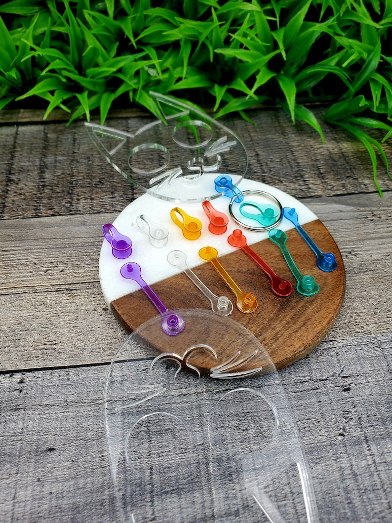 May include: A set of colorful plastic keychains in the shape of a cat. The keychains are arranged on a wooden and white circular board. The keychains are in various colors, including purple, blue, green, orange, yellow, and red.