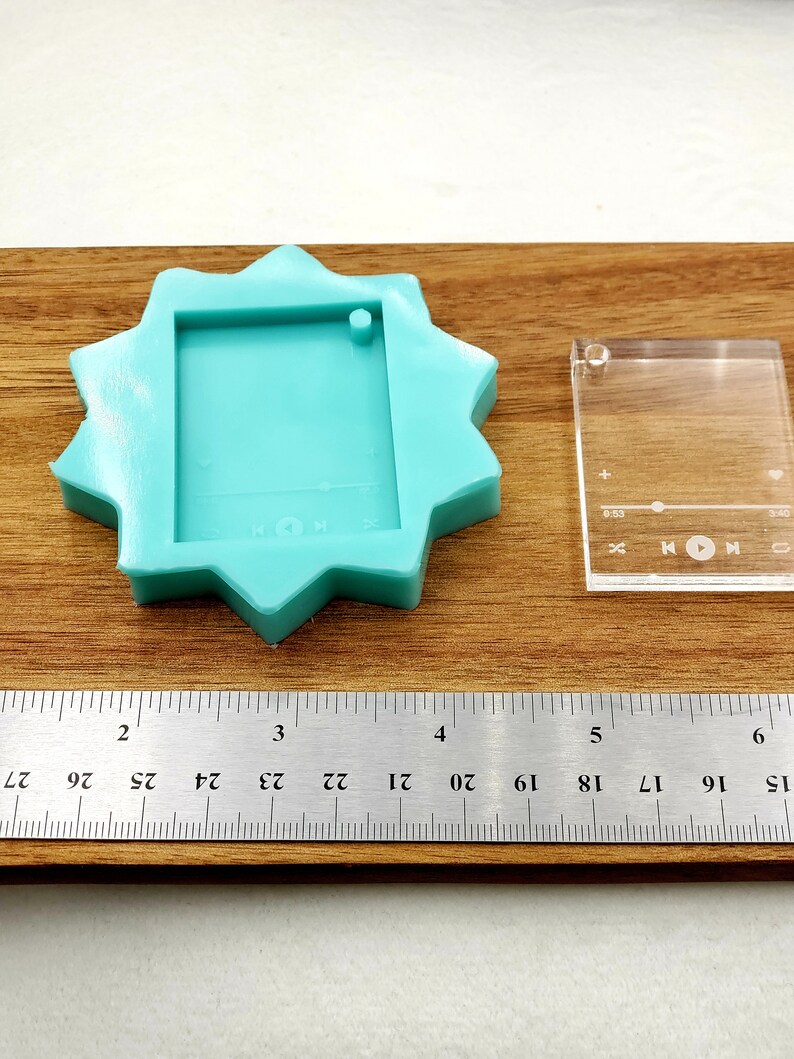 Puede incluir: Un molde de silicona turquesa con forma de estrella con un recorte rectangular en el centro. Tambi&eacute;n se muestra una plantilla de pl&aacute;stico transparente con un recorte rectangular y el texto "HON". Se puede ver una regla en la parte inferior de la imagen.