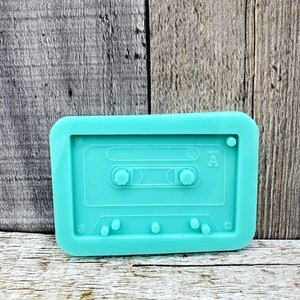 May include: A teal blue silicone mold shaped like a cassette tape. The mold has a raised design of a cassette tape with the words "side A" on the right side.