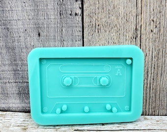 May include: A teal blue silicone mold shaped like a cassette tape. The mold has a raised design of a cassette tape with the words "side A" on the right side.