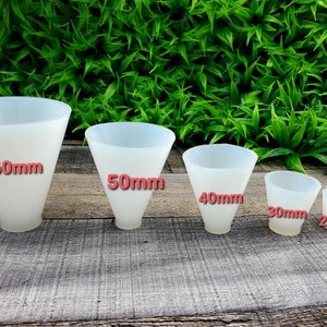 Puede incluir: Un conjunto de seis embudos de silicona blancos translúcidos de diferentes tamaños, que van de 20 mm a 60 mm de diámetro. Los embudos están dispuestos en fila, el más grande a la izquierda y el más pequeño a la derecha. Fondo de madera y follaje verde.