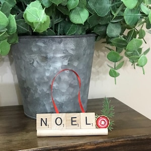 May include: A wooden block sign spells "NOEL" with Scrabble tiles, featuring a red ribbon for hanging. A small red and gold ornament with greenery accents the sign, set against a galvanized metal bucket with green foliage.