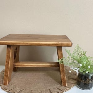 May include: A small, wooden step stool with a rectangular top and angled legs. The stool is made of light brown wood with a heart-shaped cutout on the side. A decorative plant in a glass jar sits nearby.