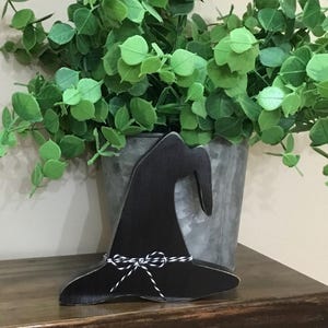 May include: A black wooden witch hat decoration with a white and black twine bow. The hat is displayed on a dark wooden surface, with a galvanized metal bucket filled with green foliage in the background.