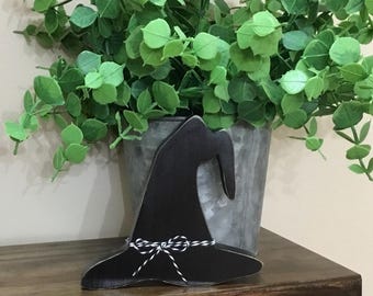 May include: A black wooden witch hat decoration with a white and black twine bow. The hat is displayed on a dark wooden surface, with a galvanized metal bucket filled with green foliage in the background.