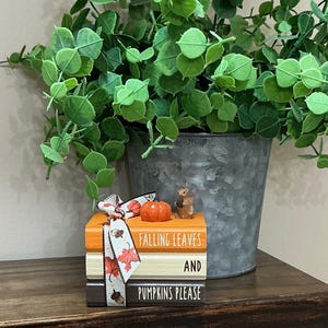 May include: A stack of three decorative books in orange, cream, and dark brown, tied with a ribbon featuring autumn leaf and acorn patterns. The books are inscribed with the text FALLING LEAVES AND PUMPKINS PLEASE, topped with a small pumpkin.