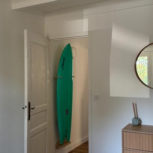 May include: A teal surfboard is mounted vertically on a wall, held by wooden supports. The board has a pointed nose and a rounded tail. The word "Stewart" is visible on the board. The room has white walls and a wooden floor.