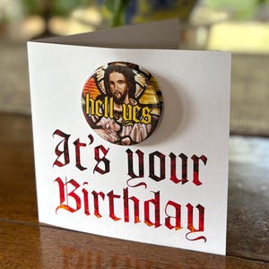 May include: A white birthday card with the text "It's your Birthday" in red and black gothic lettering. A round button with the words "hell yes" and an image of Jesus is attached to the card.