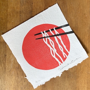 Può includere: Un foglio quadrato di carta bianca ruvida con un grande cerchio rosso al centro. Delle bacchette nere sono posizionate orizzontalmente sopra il cerchio, sollevando linee bianche ondulate che ricordano dei noodles. Firmato e numerato 2/10.