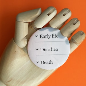May include: A white round button with black text that reads "Early life", "Diarrhea", and "Death". The button is held in a wooden hand.