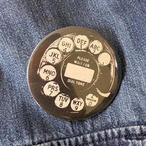 May include: A circular button with a rotary phone dial design in black and white. The button has the text "PLEASE WAIT FOR DIAL TONE" and numbers and letters. It is set against a blue denim background.