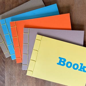 May include: A collection of handmade books with visible stitching. The covers are in grey, blue, orange, and yellow. The word "Book" is printed in blue on the yellow cover. The books are arranged on a wooden surface, with the spine stitching visible.