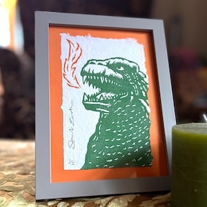 May include: Framed print of a green Godzilla illustration with orange flames and an orange background, set in a grey frame. The artwork includes the artist's signature. A green candle is in the foreground.