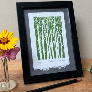 Op de afbeelding: Ingelijste kunstdruk met een witte boom-silhouet op een groene achtergrond, in een zwart frame. Het kunstwerk is gesigneerd. Een kleine vaas met gele en roze bloemen staat ernaast. Een potlood en bril zijn ook zichtbaar.