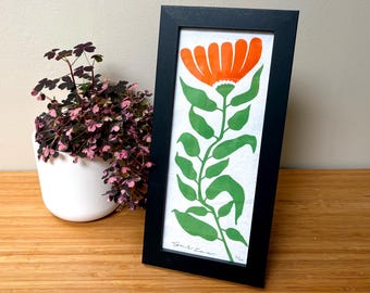 Puede incluir: Una impresión artística vertical enmarcada que presenta una flor naranja estilizada con un tallo y hojas verdes sobre un fondo blanco. La obra de arte está encerrada en un marco rectangular negro simple e incluye una firma manuscrita y número de edición.