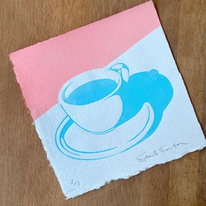 May include: A print depicting a blue teacup and saucer on a white background, set against a pink backdrop. The artwork has a minimalist style with the artist's signature and the number 2/5.