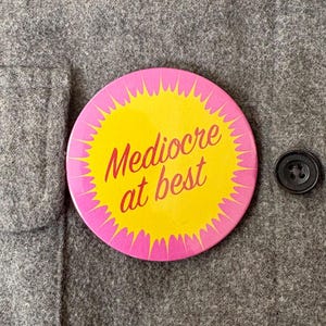 May include: A round button with the text "Mediocre at best" in red on a yellow background, framed by a pink burst design. The button is pinned to a gray fabric, next to a black button.
