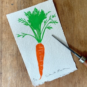 May include: A rectangular piece of off-white textured paper featuring a hand-printed orange carrot with vibrant green leafy tops. The print is signed and numbered 1/10 in the bottom corner, accompanied by a wooden-handled metal carving tool.