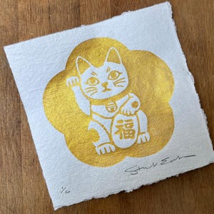 May include: A square piece of white textured paper featuring a gold foil print of a beckoning cat centred within a gold flower shape. The cat holds an oval with the Chinese character for luck. The print is signed and numbered 1/6 in black ink.