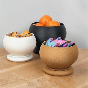 May include: Three ceramic bowls, one white, one black, and one brown, with different contents. The black bowl holds oranges, the white bowl holds yellow and orange sweets, and the brown bowl holds blue and pink sweets.