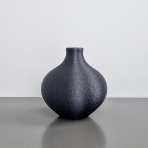 Könnte beinhalten: Eine schwarze Keramikvase mit runder, bauchiger Form. Die Vase hat eine strukturierte Oberfläche und einen glatten, abgerundeten Rand.