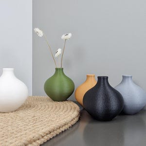 Könnte beinhalten: Fünf kleine, strukturierte Vasen in Weiß, Grün, Orange, Schwarz und Hellblau. Sie stehen auf einer gewebten Matte und einer grauen Oberfläche. Eine Vase enthält ein paar weiße Blumen.