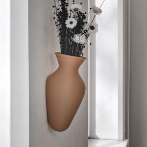 Peut inclure: Un vase en céramique beige avec une surface texturée, monté sur un mur blanc, contient un bouquet de fleurs blanches séchées avec des tiges brun foncé.
