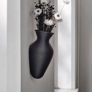 May include: A black ceramic vase with a textured finish, mounted on a white wall, holds a bouquet of dried white daisies.