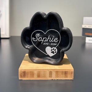 May include: A black paw-shaped memorial with the name "Sophie" and dates 2012-2024 in a heart-shaped design. The memorial sits on a wooden base, suitable for pet remembrance.