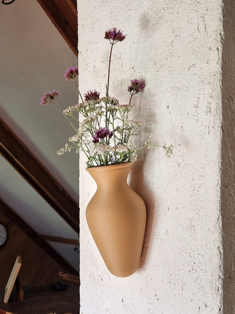 May include: A tan ceramic vase with a textured surface is mounted on a white wall. The vase is filled with a bouquet of pink and white flowers.