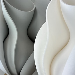 Op de afbeelding: Twee abstracte sculpturen met organische vormen. Eén is lichtgrijs, de andere wit. De sculpturen hebben een gelaagd, bloembladachtig ontwerp, met gladde, gebogen oppervlakken. De sculpturen staan op een houten voet.