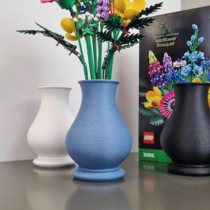 Op de afbeelding: Een set van blauwe, witte en zwarte keramische vazen met een boeket kleurrijke LEGO-bloemen. De doos van de LEGO Wildflower Bouquet set is zichtbaar op de achtergrond.